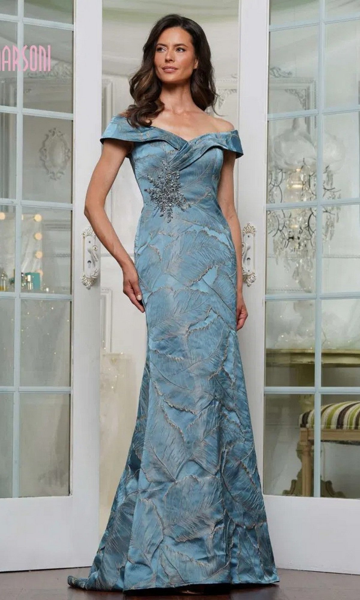 A woman stands indoors wearing the Marsoni MV1266 long formal dress in teal, an off-the-shoulder evening gown with a floor-length design, leaf patterns, and a decorative waist embellishment.